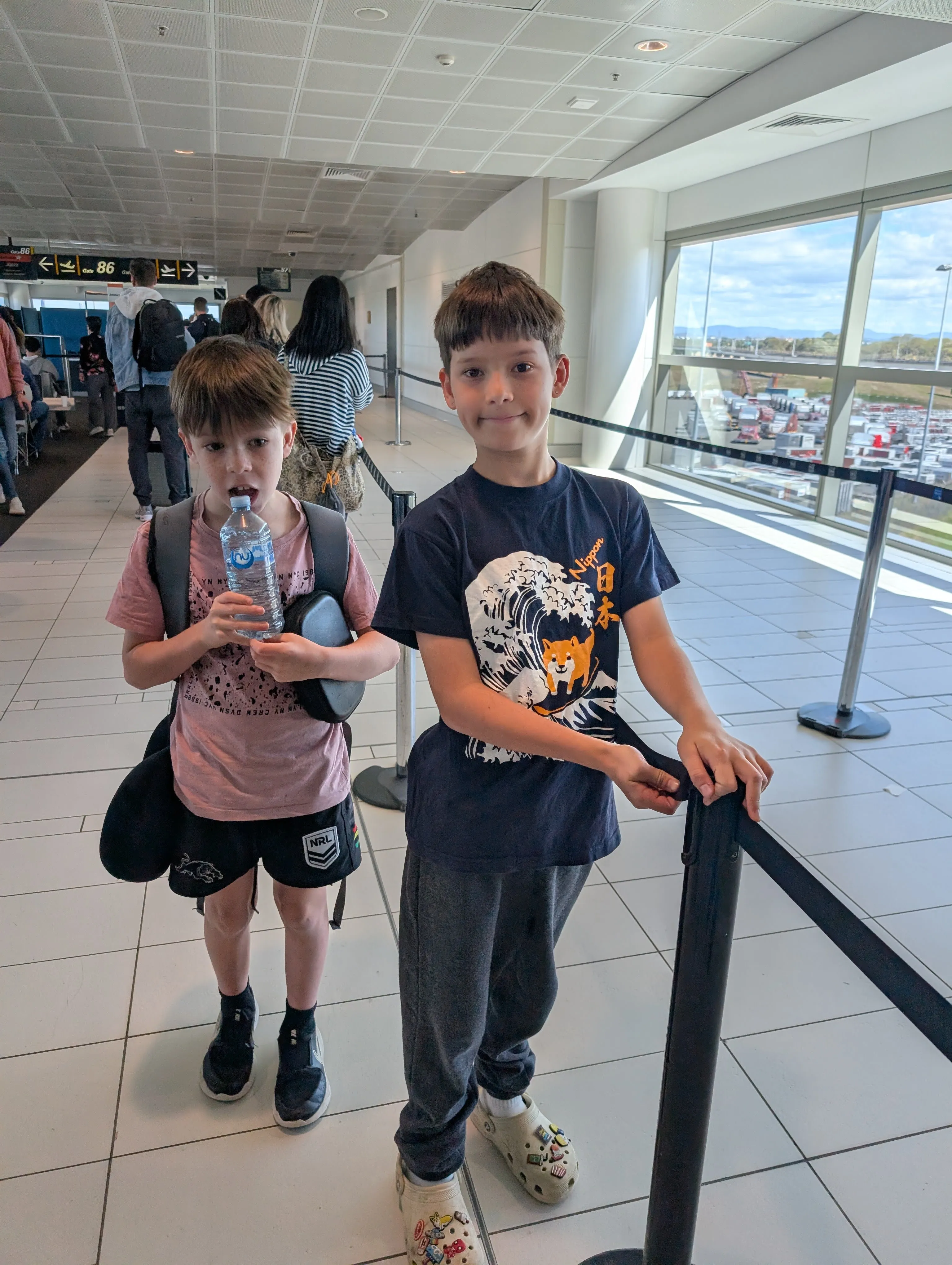 The two boys at the departure gate with carry-on backpacks, a water bottle and a neck pillow, one in a Japan wave t-shirt