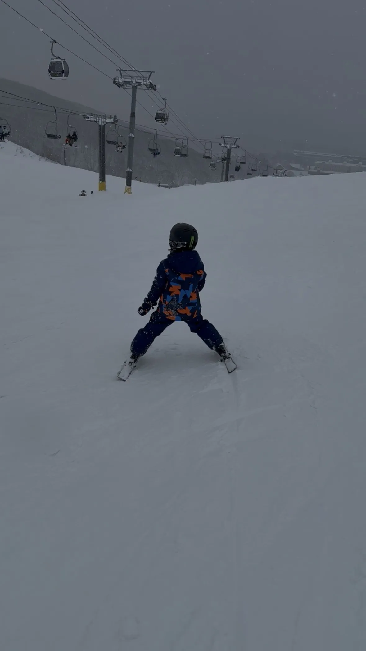 A young child in a camo ski suit skiing down a wide open run at Grand Hirafu, with the gondola and chairlifts above