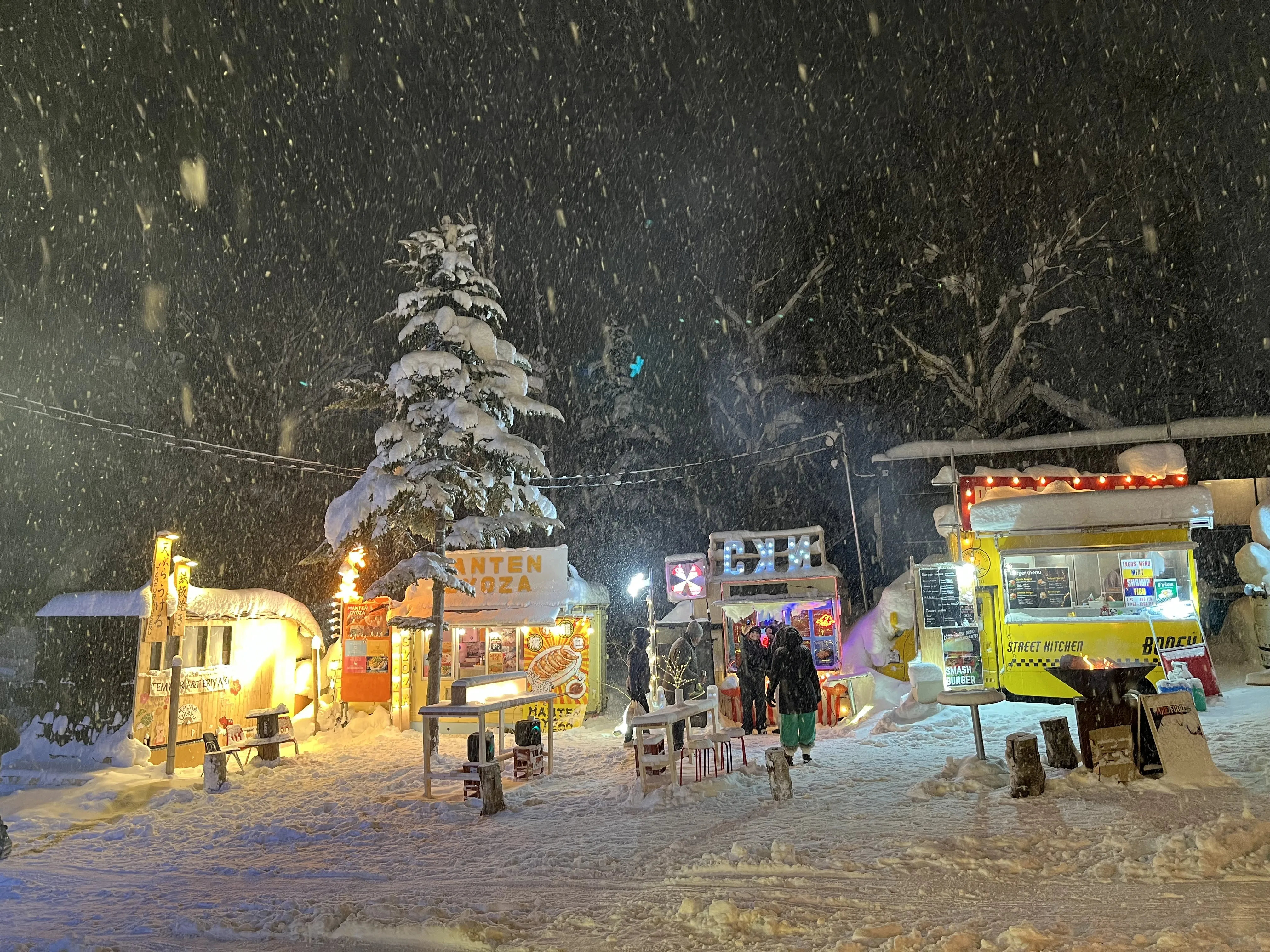 Brightly lit food stalls and a food-truck glowing in heavy snowfall at night in Niseko Hirafu