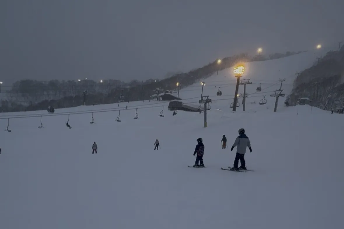 Skiers coming down a floodlit Grand Hirafu run at night with the village lights below